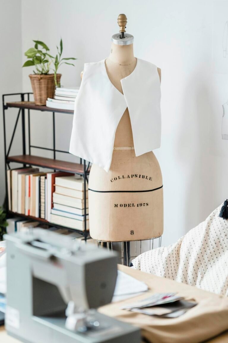 Table with professional sewing machine placed near mannequin with white cloth near wall in light modern atelier with collection of books